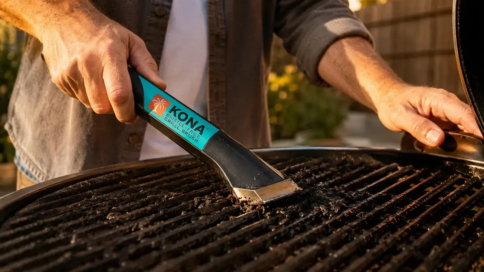 Hand gripping the Kona bristle-free grill brush handle and scrubbing a cast iron grate with visible grease residue being removed
