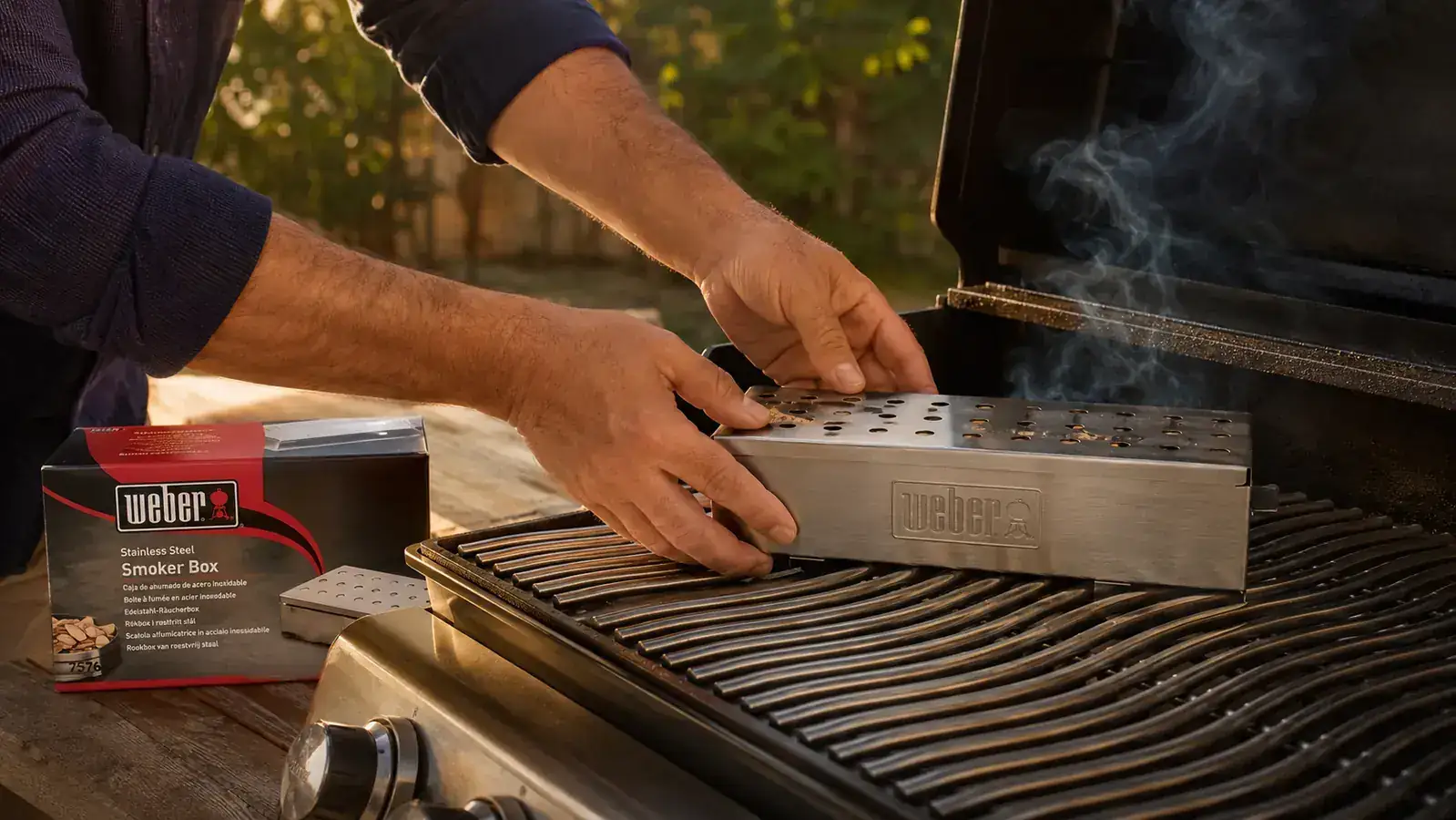 Hand filling the Weber Premium Smoker Box with hickory wood chips, hinged lid open, sitting on gas grill grates